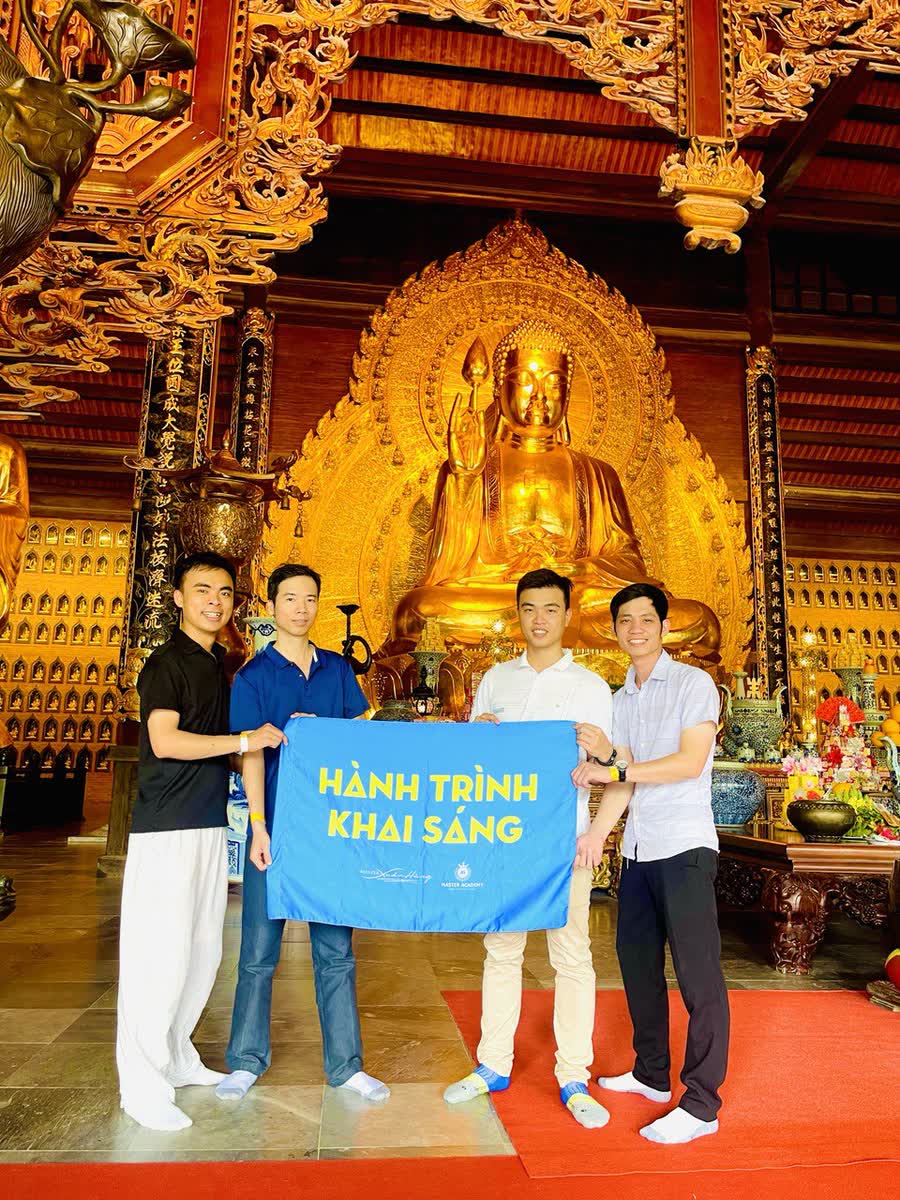 Bốn người cầm lá cờ Hành Trình Khai Sáng trong một chánh điện, trước tượng Phật lớn